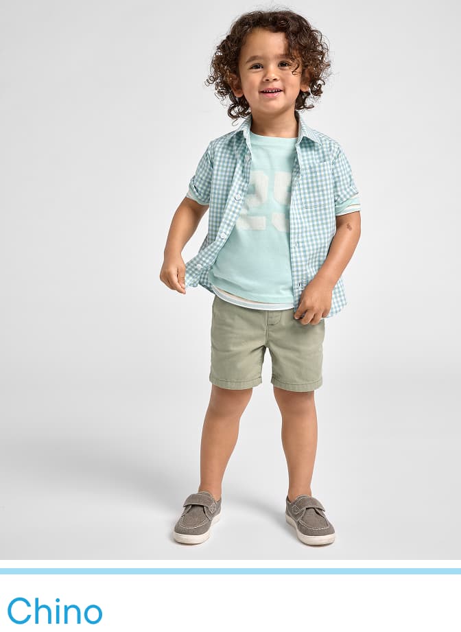 A young child with curly hair stands in a light-colored outfit, smiling against a plain white background.