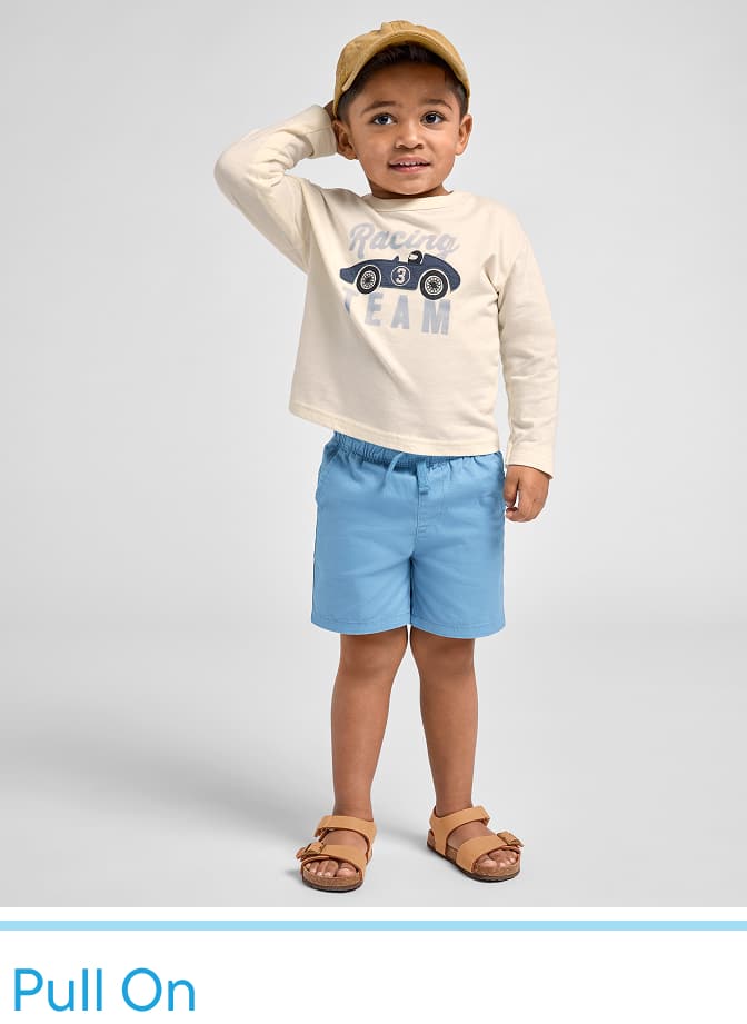 A young boy wearing a beige shirt with a car graphic, blue shorts, and sandals stands against a plain white background, smiling and gesturing with his hand.