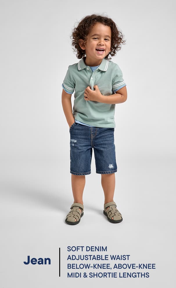 Un niño pequeño con cabello rizado está de pie y sonriendo, vistiendo una camiseta polo verde claro y pantalones cortos de mezclilla, sobre un fondo blanco liso.