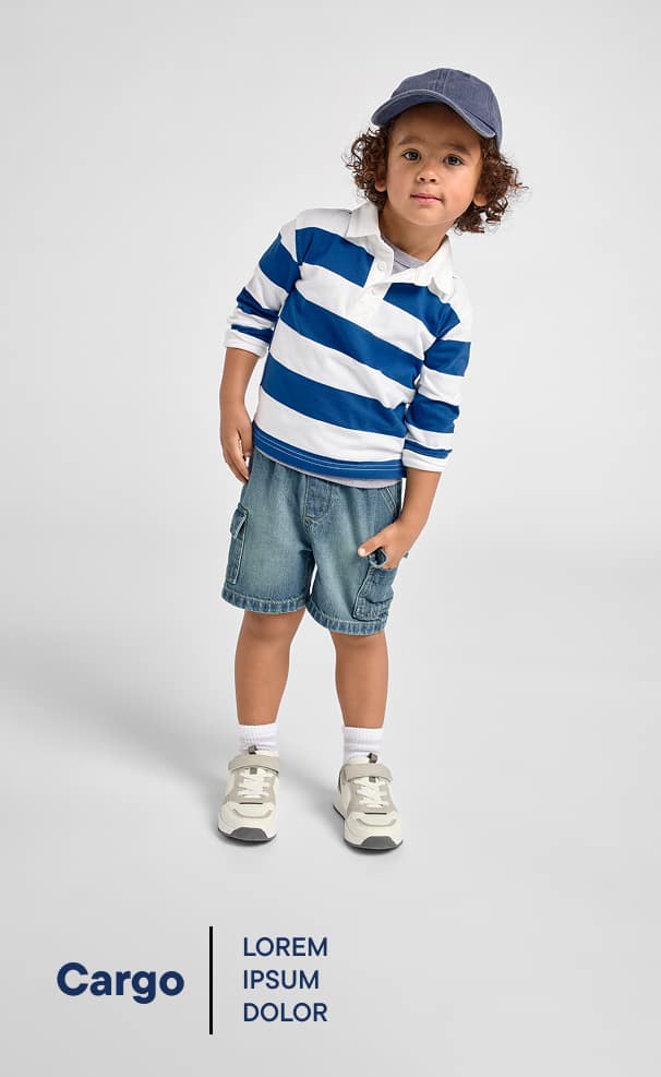 Un niño pequeño, vestido con una camiseta de rayas, pantalones cortos vaqueros y zapatillas deportivas, aparece de pie sobre un fondo blanco liso, con una expresión alegre en el rostro.