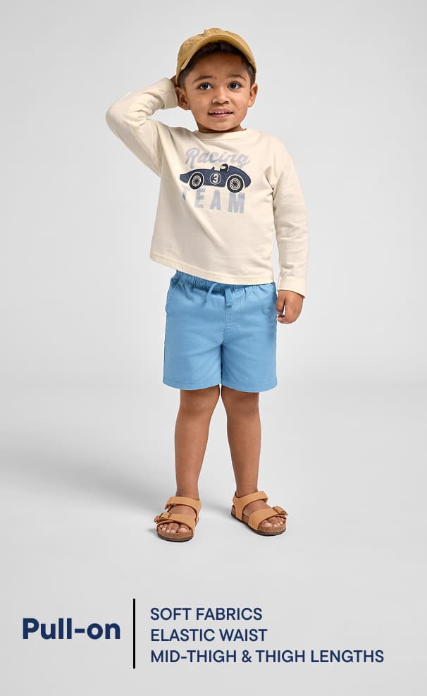 Un niño pequeño, vestido con una camiseta beige con el dibujo de un coche, pantalones cortos azules y sandalias, aparece de pie sobre un fondo blanco liso, sonriendo y gesticulando con la mano.