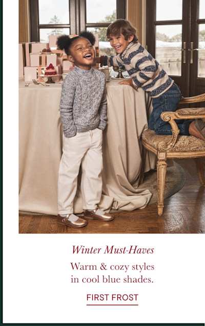 A young girl with curly hair is smiling and wearing a striped sweater with penguin designs, standing in front of a stone wall.