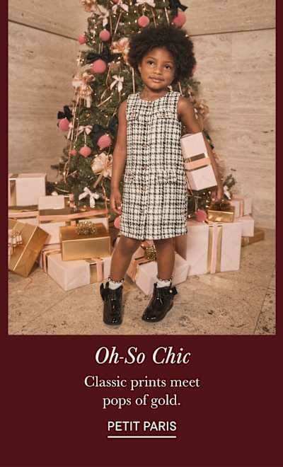 A young child with curly hair stands in front of a Christmas tree, surrounded by wrapped gifts, wearing a checkered dress and black shoes.