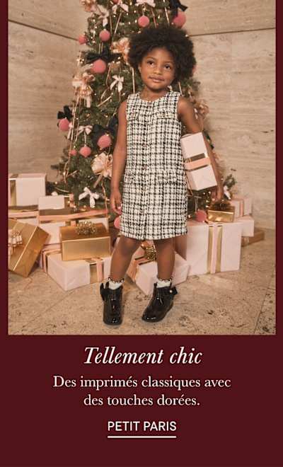 Une jeune enfant aux cheveux bouclés se tient devant un sapin de Noël, entourée de cadeaux emballés, vêtue d'une robe à carreaux et de chaussures noires.