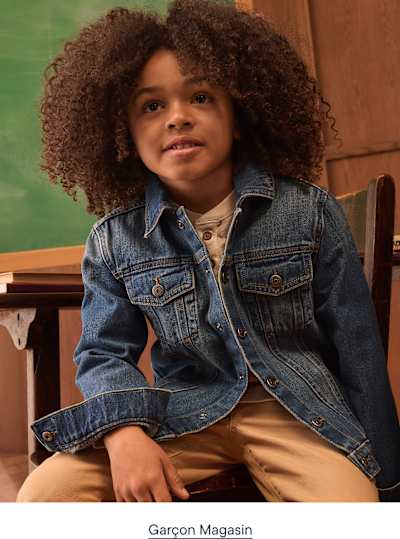 Un jeune garçon aux cheveux bouclés, vêtu d'une veste en jean et d'un pantalon beige, est assis devant un tableau noir.