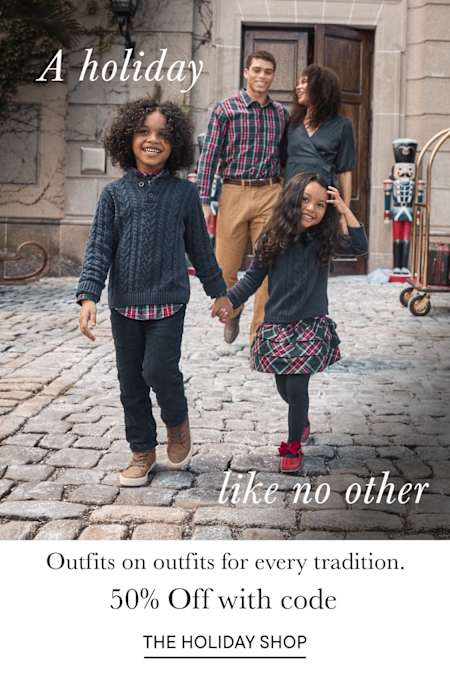 The image shows a group of four young people, two men and two women, walking together down a cobblestone street lined with buildings and flowers. They appear to be dressed in casual, stylish outfits, suggesting a holiday or vacation setting.