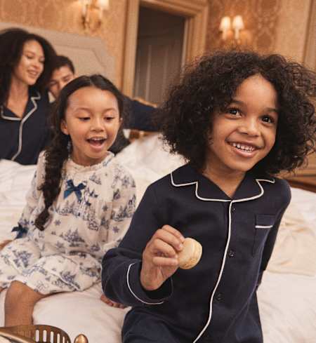 The image shows two young girls, one with curly hair and the other with straight hair, sitting on a couch and smiling happily. The background appears to be a cozy, well-lit room with decorative elements.