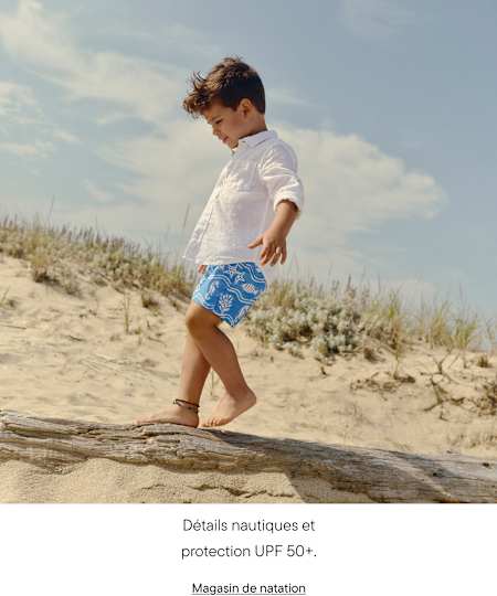 Un jeune enfant vêtu de blanc et d'un short bleu à motifs marche le long d'un tronc d'arbre sur une plage de sable, sous un ciel nuageux et avec de la végétation en arrière-plan.