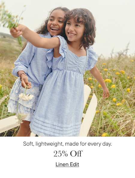 Two young girls in blue dresses are embracing and smiling joyfully in a field of wildflowers, with a wooden fence visible in the background.