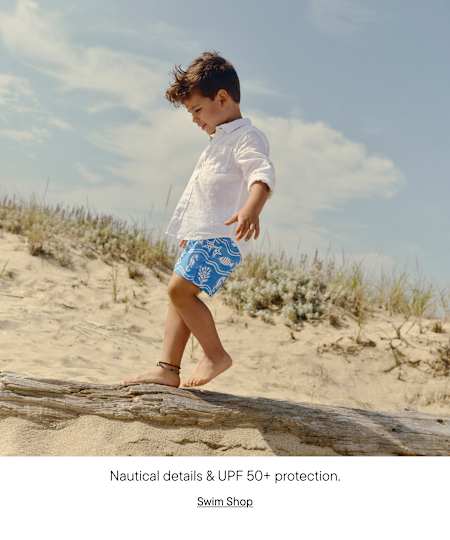 A young child in casual clothing walks along a wooden log on a sandy beach, with a cloudy sky and vegetation in the background.
