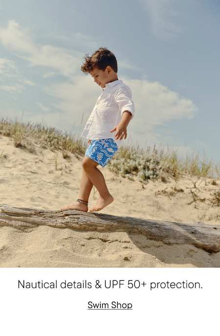 A young child in white clothing and blue patterned shorts walks along a sandy beach with dunes and a cloudy sky in the background.