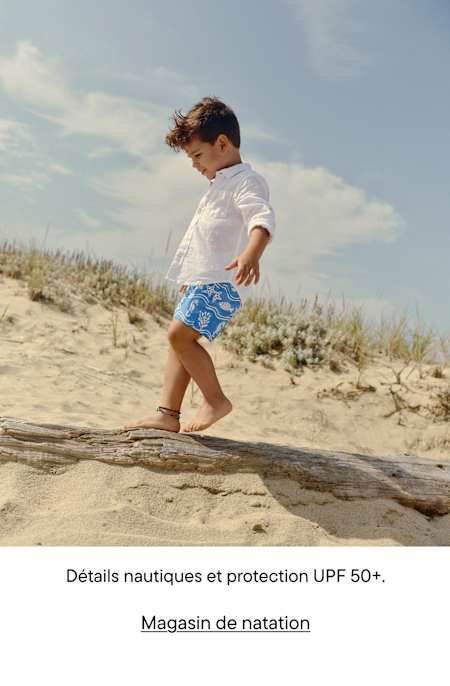 Un jeune enfant vêtu de blanc et d'un short bleu à motifs marche le long d'une plage de sable fin, avec des dunes et un ciel nuageux en arrière-plan.