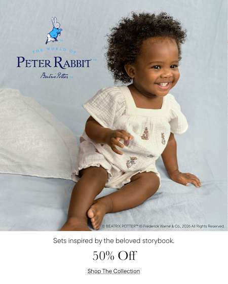 A smiling young child with curly hair sits on a white surface, wearing a white outfit with a Peter Rabbit logo, against a light blue background.