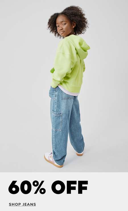 A young person with curly hair wearing a bright green puffy jacket and loose-fitting jeans stands against a plain white background, with a "60% OFF SHOP JEANS" text overlay.