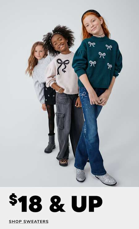 Three young girls wearing stylish sweaters and jeans stand together in a studio setting, with a promotional message for a sweater sale displayed in the background.