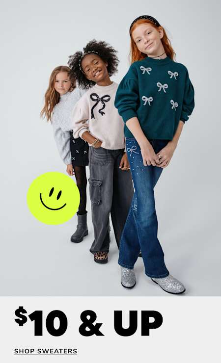 Three young girls wearing stylish sweaters and jeans stand together in a studio setting, with a promotional message for a sweater sale displayed in the background.