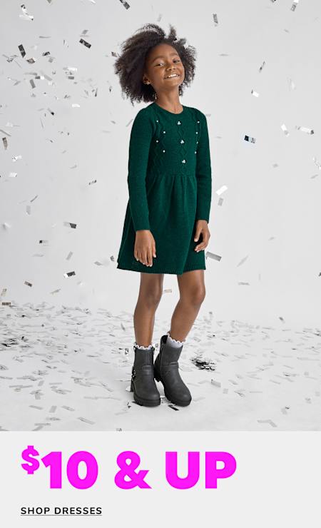 A young girl wearing a green dress and boots stands in a snowy setting, surrounded by falling confetti-like pieces.