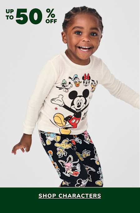 A young child wearing a white long-sleeved shirt with a Mickey Mouse design and black pants with a colorful cartoon pattern, standing against a plain white background with a green banner advertising a sale on character-themed merchandise.