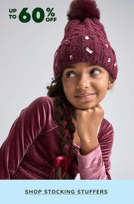 A young woman wearing a burgundy knit hat with a fur pom-pom, smiling at the camera against a background with a "Up to 60% off" sale sign for "Stocking Stuffers".