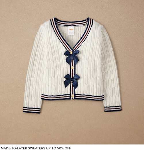 A white cable-knit cardigan with navy blue trim and decorative bows, displayed against a beige background.