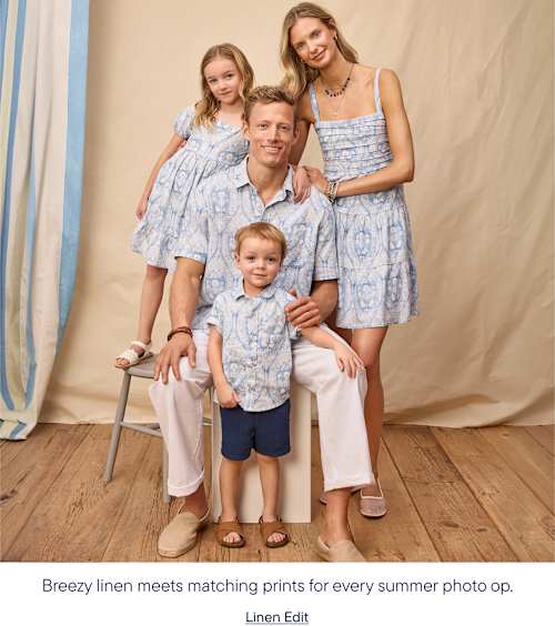 A family of four, dressed in matching blue and white patterned outfits, poses in front of a light-colored, draped backdrop with vertical striped curtains on the left.