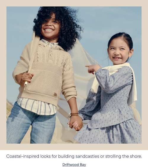 Two smiling children, a boy with curly hair and a girl with dark hair, hold hands on a sandy beach with a light blue sky in the background.