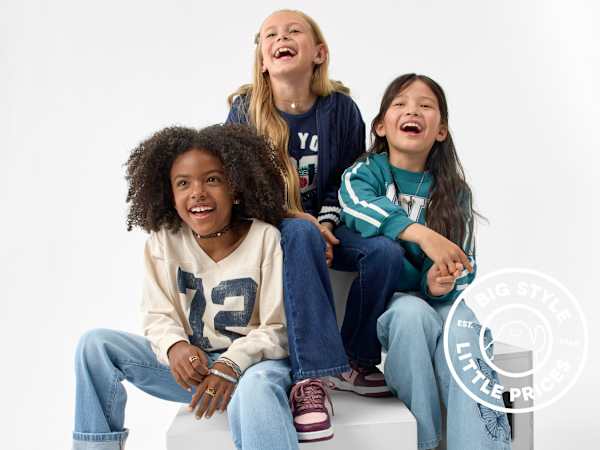 Three laughing young girls in casual clothing sit together against a plain white background.