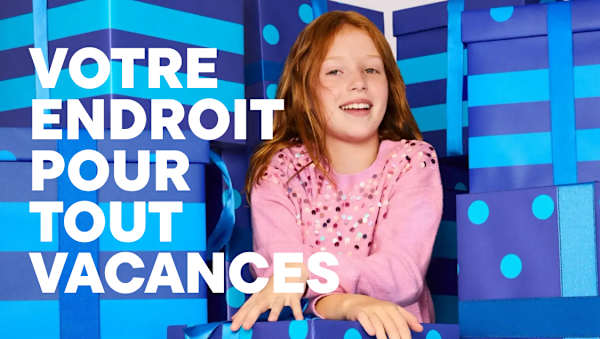 Au premier plan, une jeune fille souriante vêtue d'un pull rose est assise, entourée en arrière-plan de grands paquets cadeaux bleus et violets.