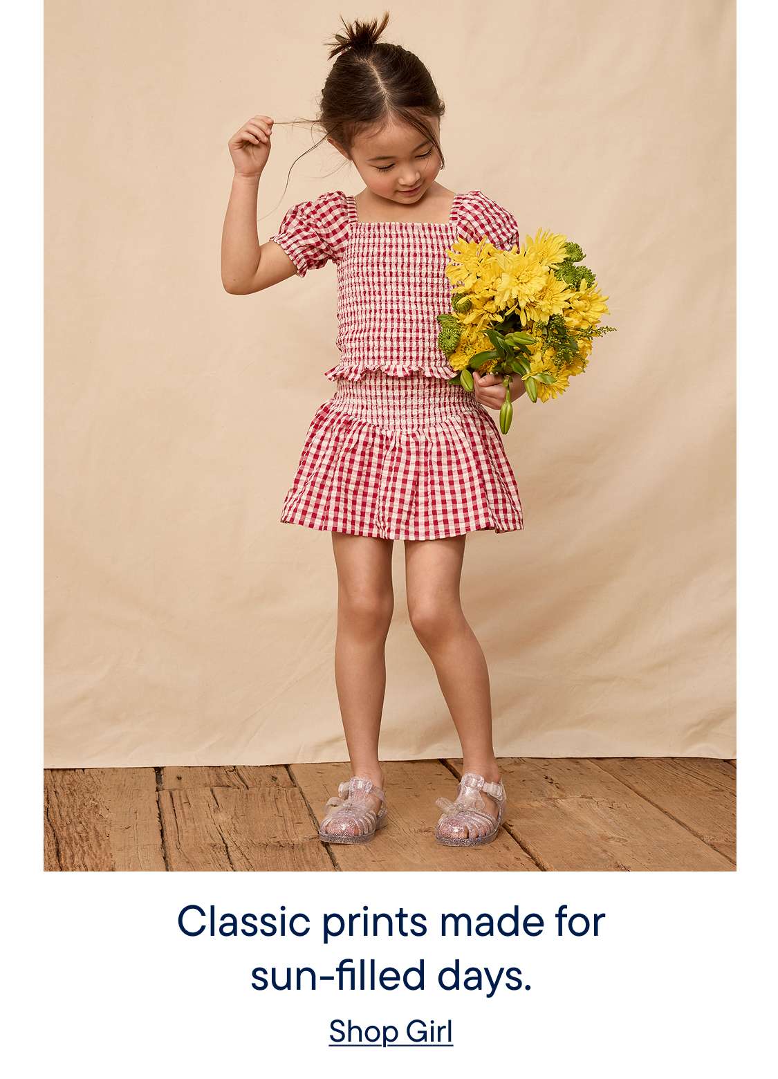 A young girl in a red and white gingham dress stands on a wooden floor, holding a bouquet of yellow flowers against a plain beige background.