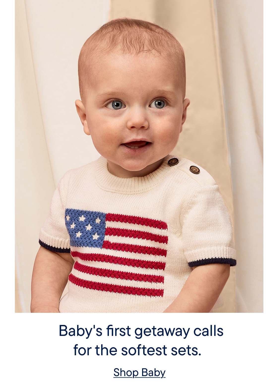 A smiling baby sits on a wooden surface, wearing a t-shirt with an American flag design, against a neutral-colored curtain backdrop.