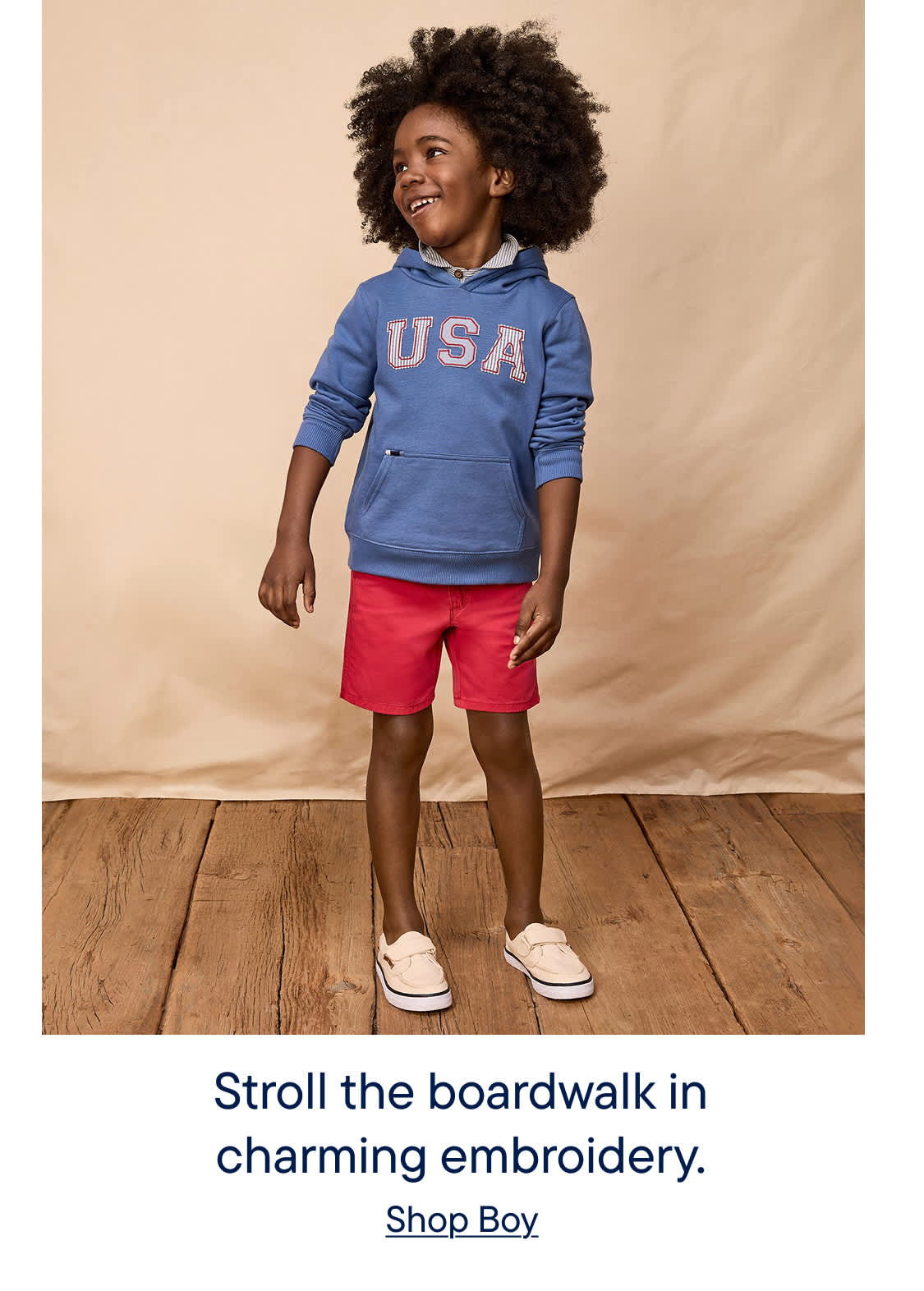 A young child with curly hair stands on a wooden floor, wearing a blue hoodie with "USA" printed on it and red shorts, exuding a charming and playful demeanor.